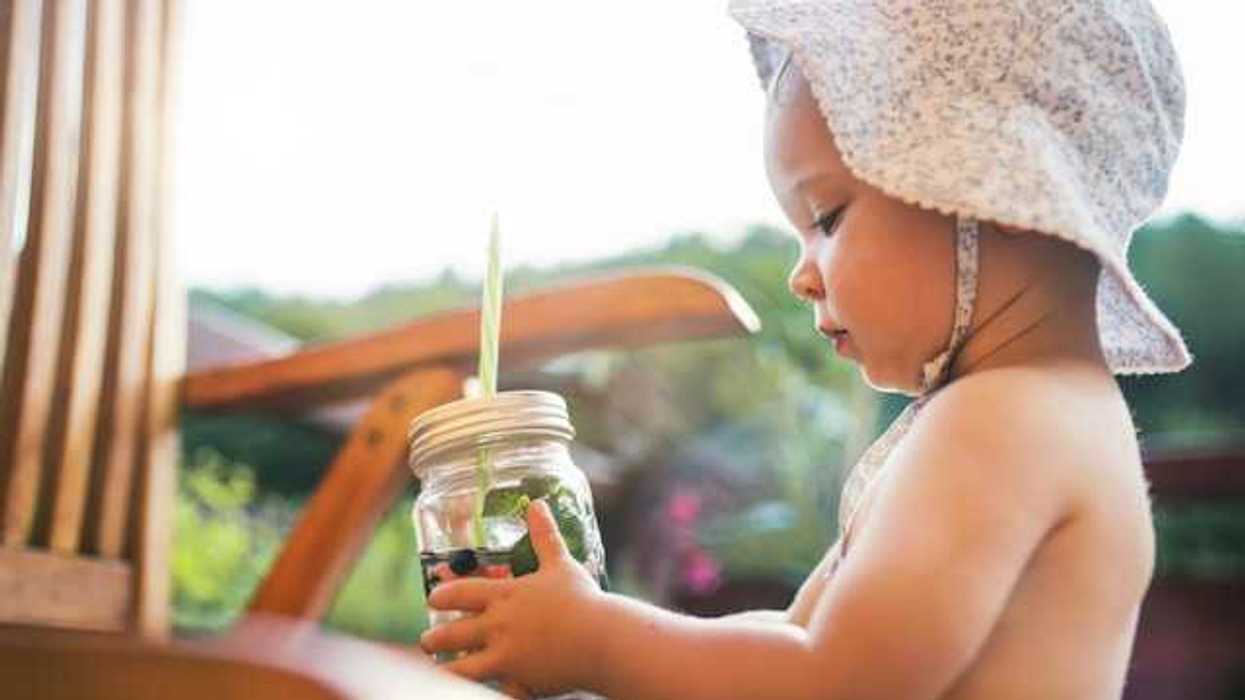 A baby with a hat on holding a glass jar with water and fruit in it