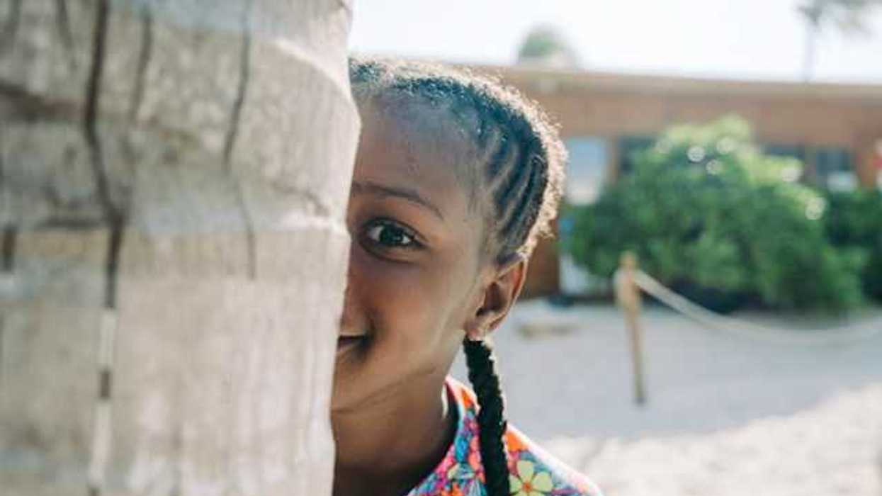 A Black child peers at the camera from behind a tree trunk