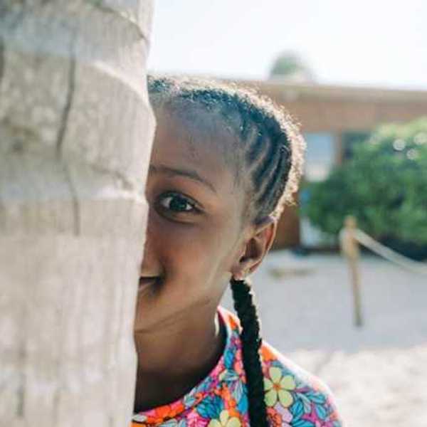 A Black child peers at the camera from behind a tree trunk