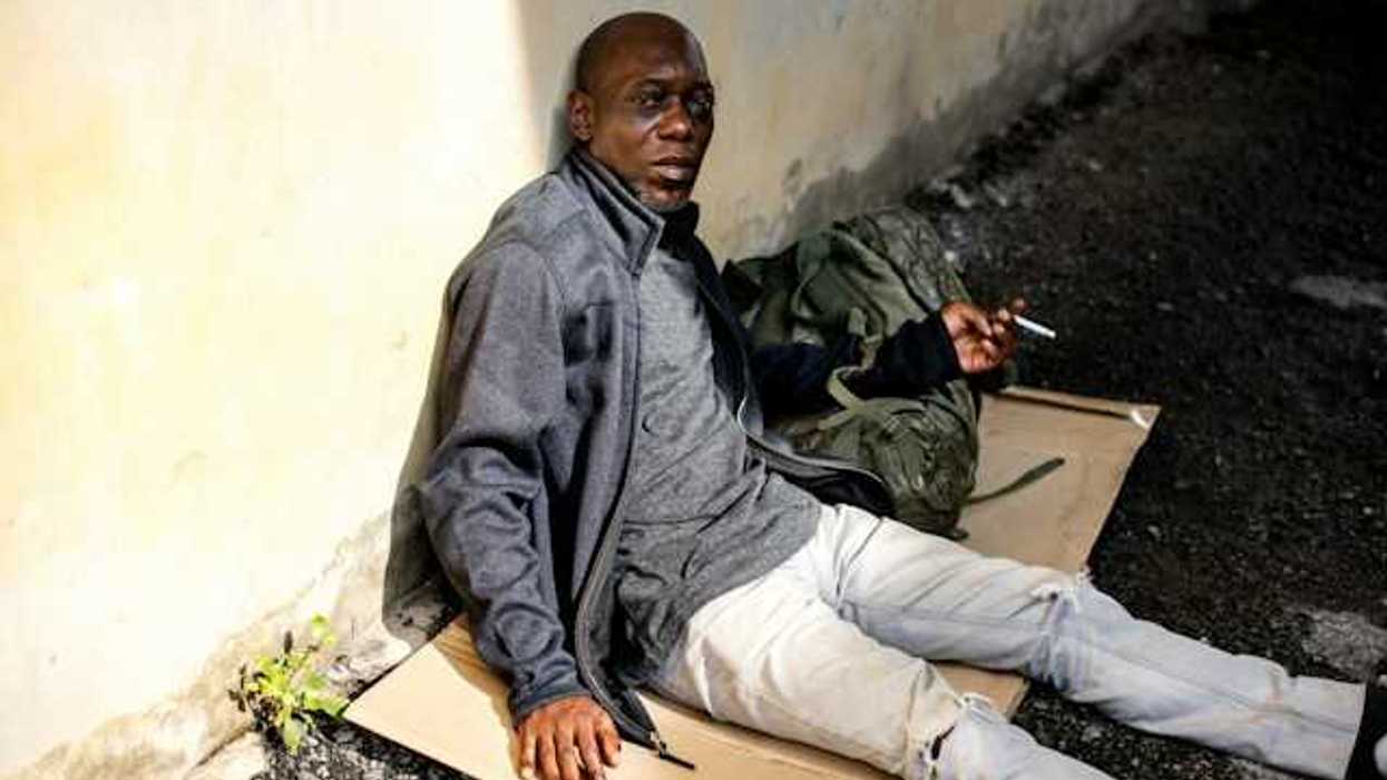 A Black homeless man sits on a piece of cardboard