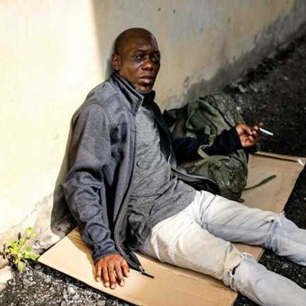 A Black homeless man sits on a piece of cardboard