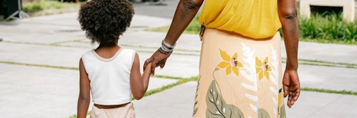 A black mother wearing a yellow top and floral print skirt holding a child's hand