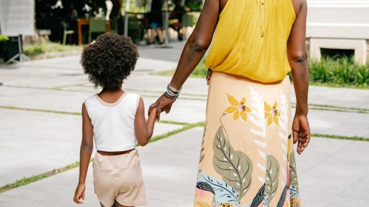 A black mother wearing a yellow top and floral print skirt holding a child's hand