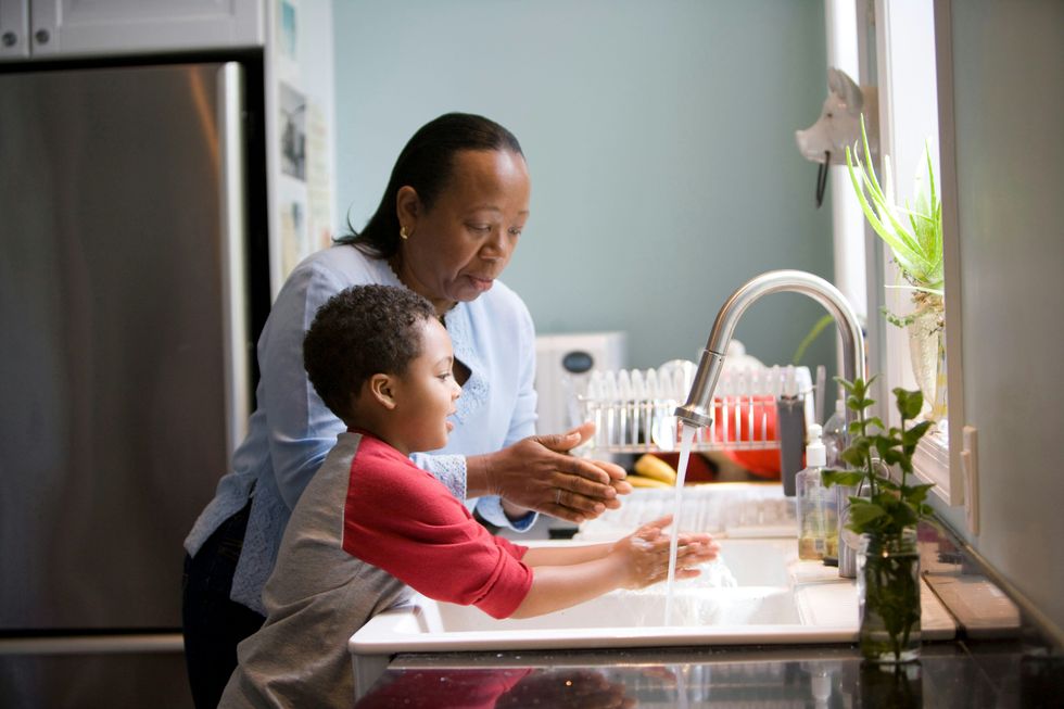 A Black woman helps a Black child wash his hands at a kitchen sink by a window.