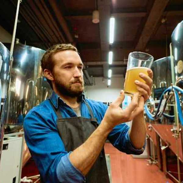 A brewer holding a glass of beer surrounded by vats