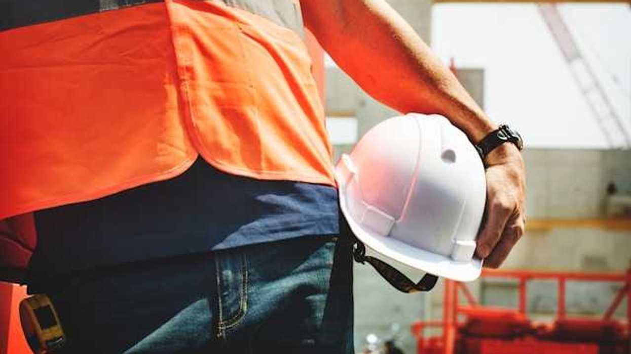 A construction worker in an orange vest carrying a white hard hat on a sunny day