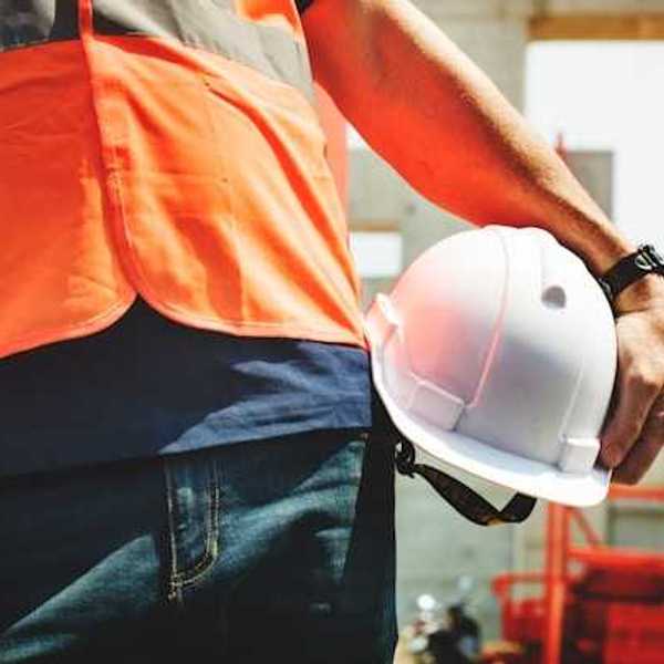 A construction worker in an orange vest carrying a white hard hat on a sunny day