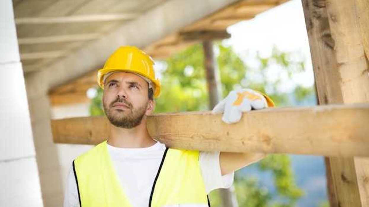 A construction worker wearing a white tshirt and a yellow safety vest carrying a piece of wood over his shoulder