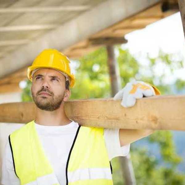 A construction worker wearing a white tshirt and a yellow safety vest carrying a piece of wood over his shoulder