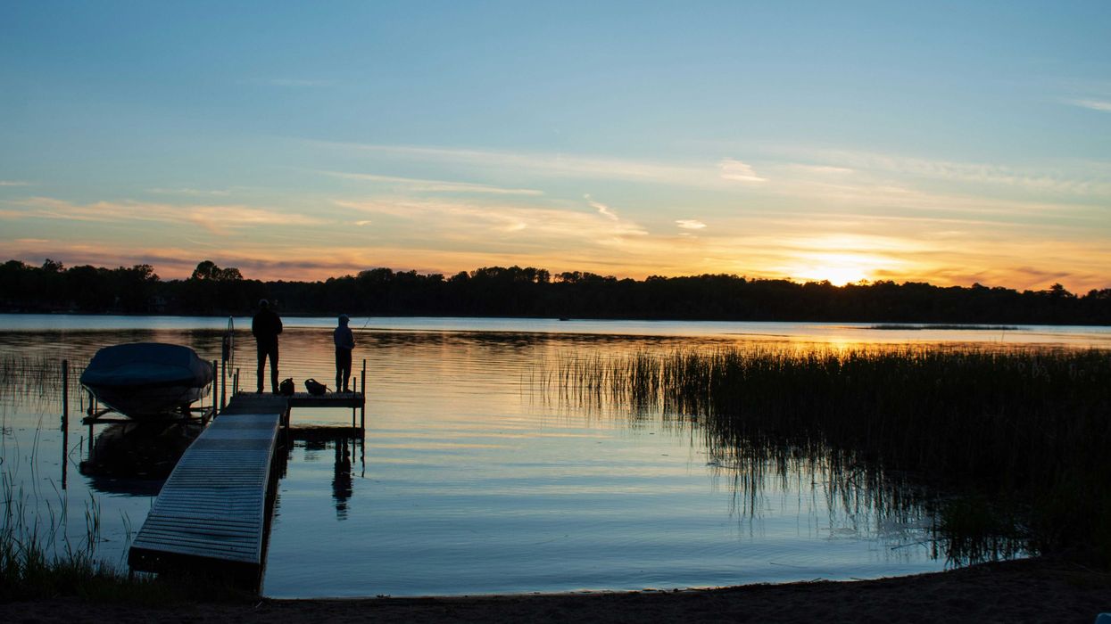 a couple of people standing on a lakeside dock at sunset