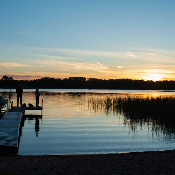a couple of people standing on a lakeside dock at sunset
