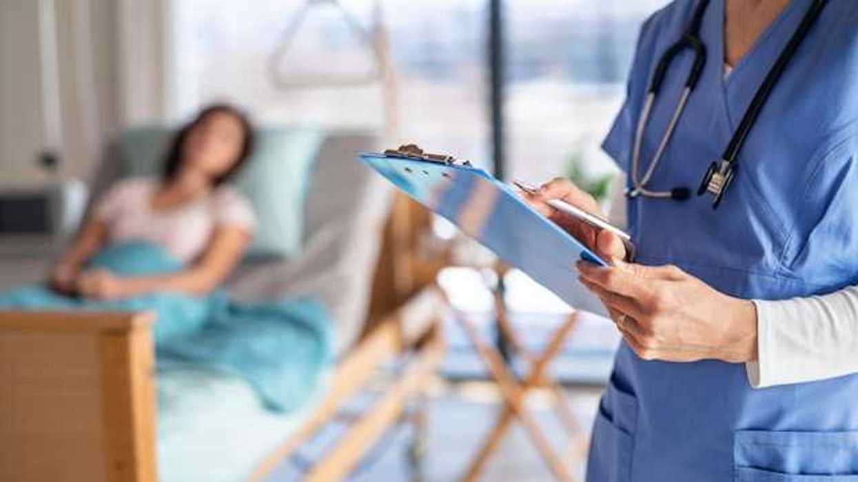 A doctor with a clipboard stands in a hospital room with a patient in a bed in the background