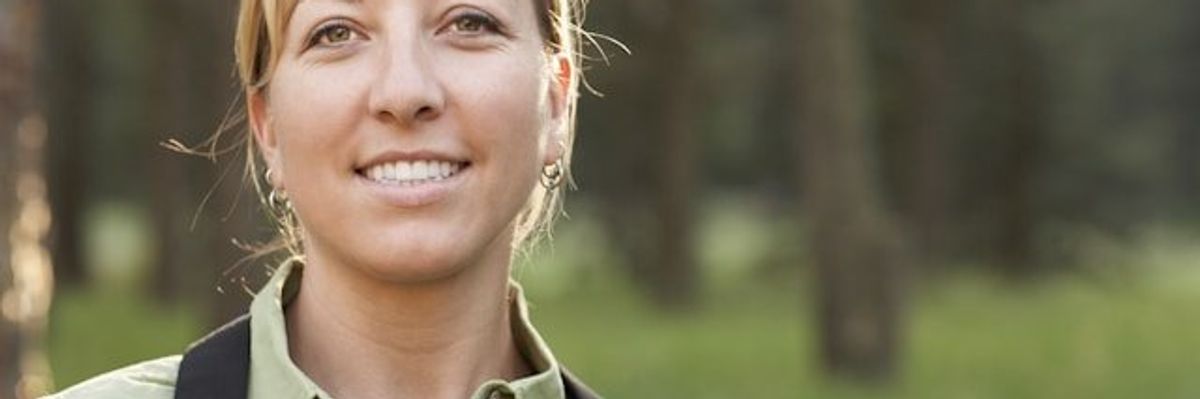 A female hiker in a green shirt with a black backpack looking into the camera.