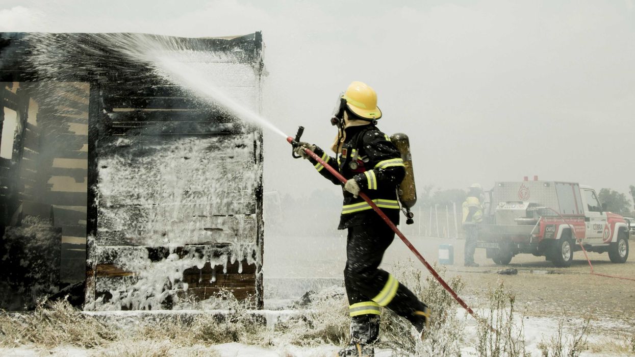 A firefighter in protective gear and helmet sprays firefighting foam on a burned building.