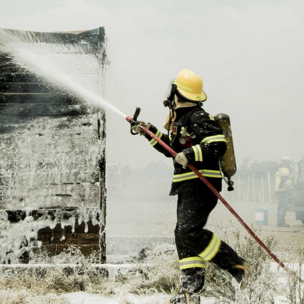 A firefighter in protective gear and helmet sprays firefighting foam on a burned building.
