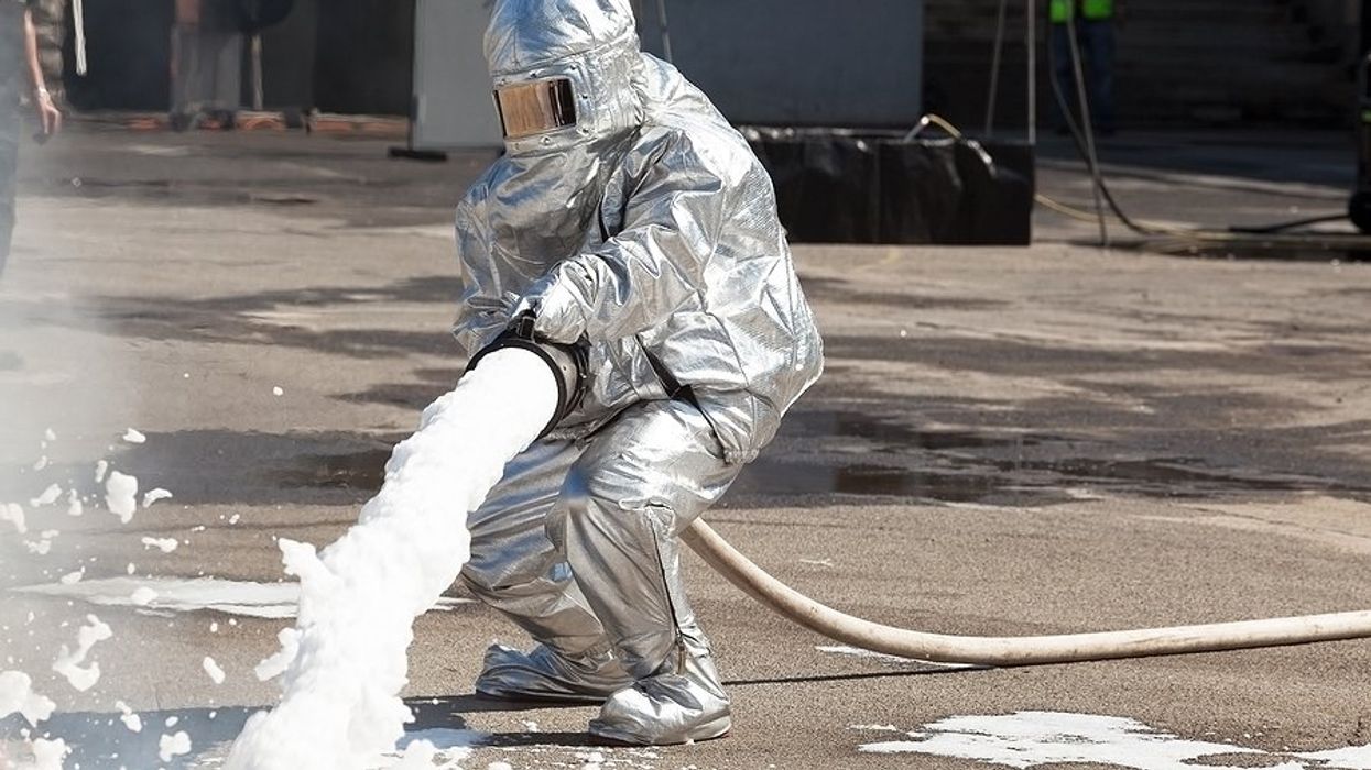 A firefighter in silver full protective gear sprays foam out of a large hose onto the ground.