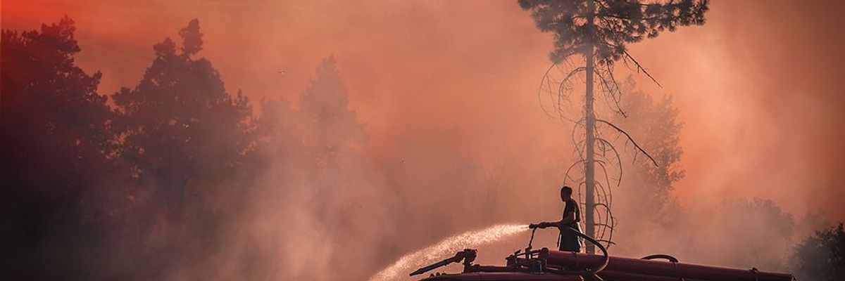 A firefighter stands on an old fire truck spraying water on grass with wildfire smoke in the air