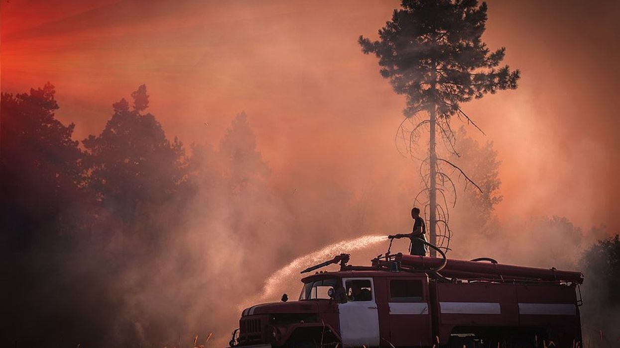 A firefighter stands on an old fire truck spraying water on grass with wildfire smoke in the air