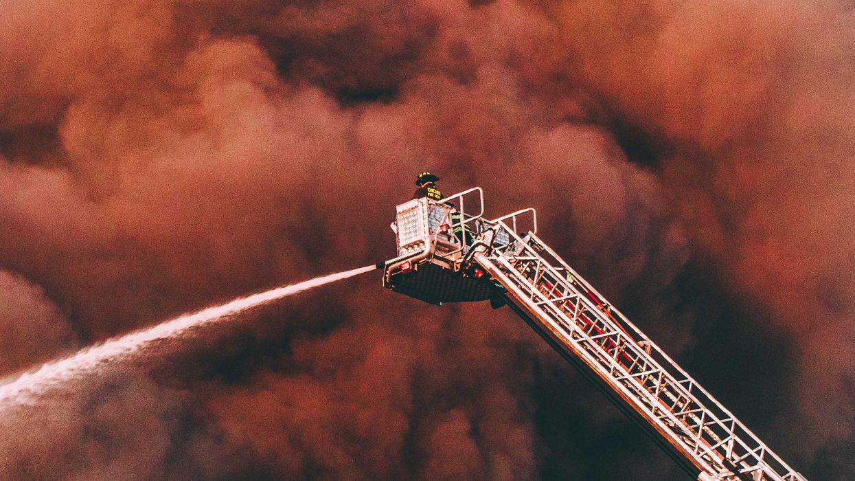 A firefighter surrounded by thick smoke sprays water on a fire from an elevated crane.