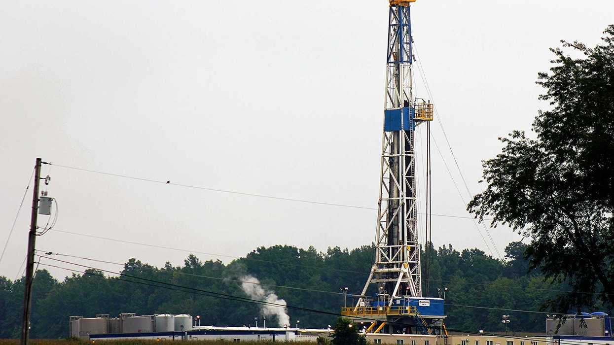 A fracking rig and infrastructure with a cornfield in foreground and trees in background.