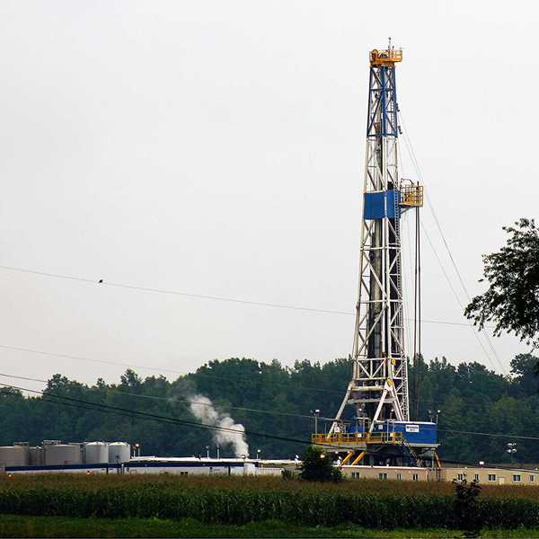 A fracking rig and infrastructure with a cornfield in foreground and trees in background.