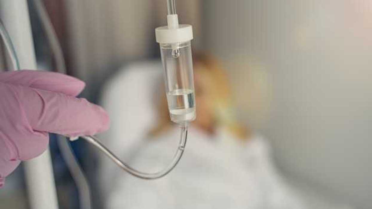 A gloved hand adjusting the plastic tube on an IV with a patient in the background