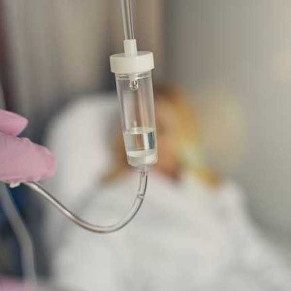 A gloved hand adjusting the plastic tube on an IV with a patient in the background