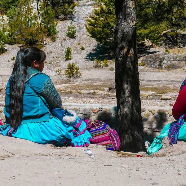 A group of Indigenous women in skirts and scarves sitting next to each other under a tree.
