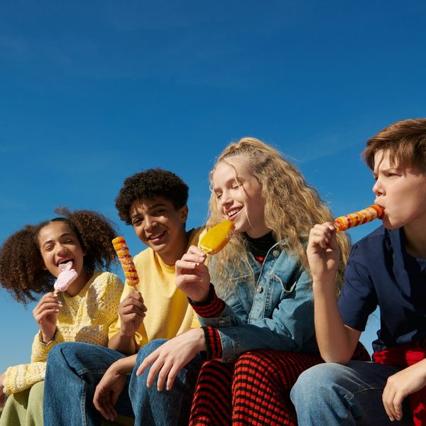 A group of teens or tweens of different races sit together eating brightly colored lollipops and ice cream on a sunny day.
