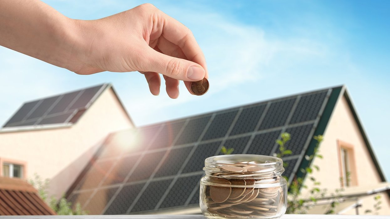 A hand places pennies in a glass jar with an image of a house roof covered in solar panels in background.