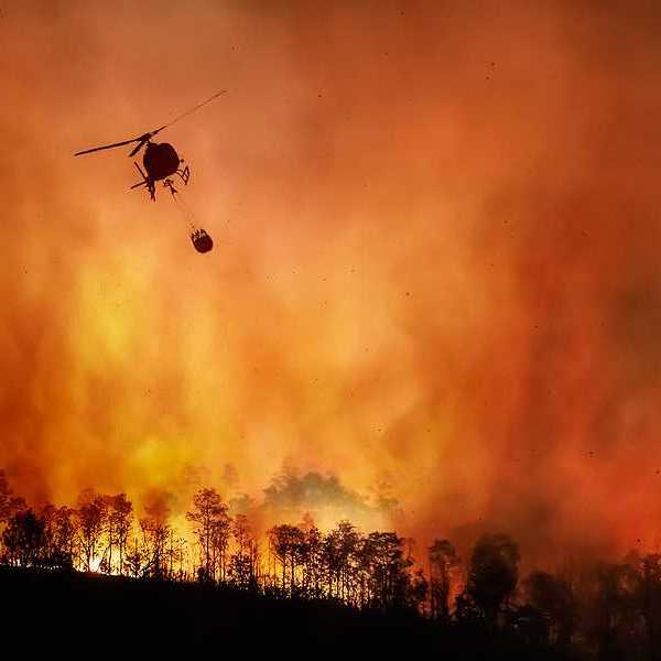 A helicopter dropping water onto a wildfire with burning trees in the background