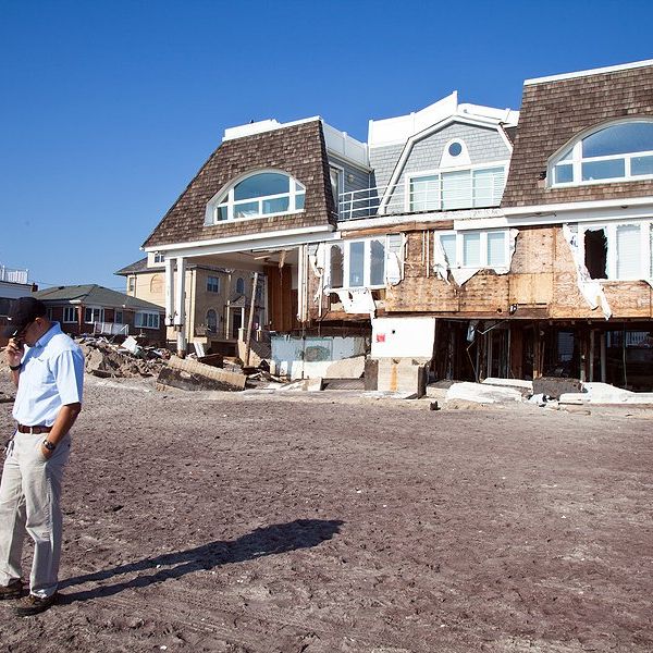 A hurricane damaged house with a man talking on the phone in front of it.