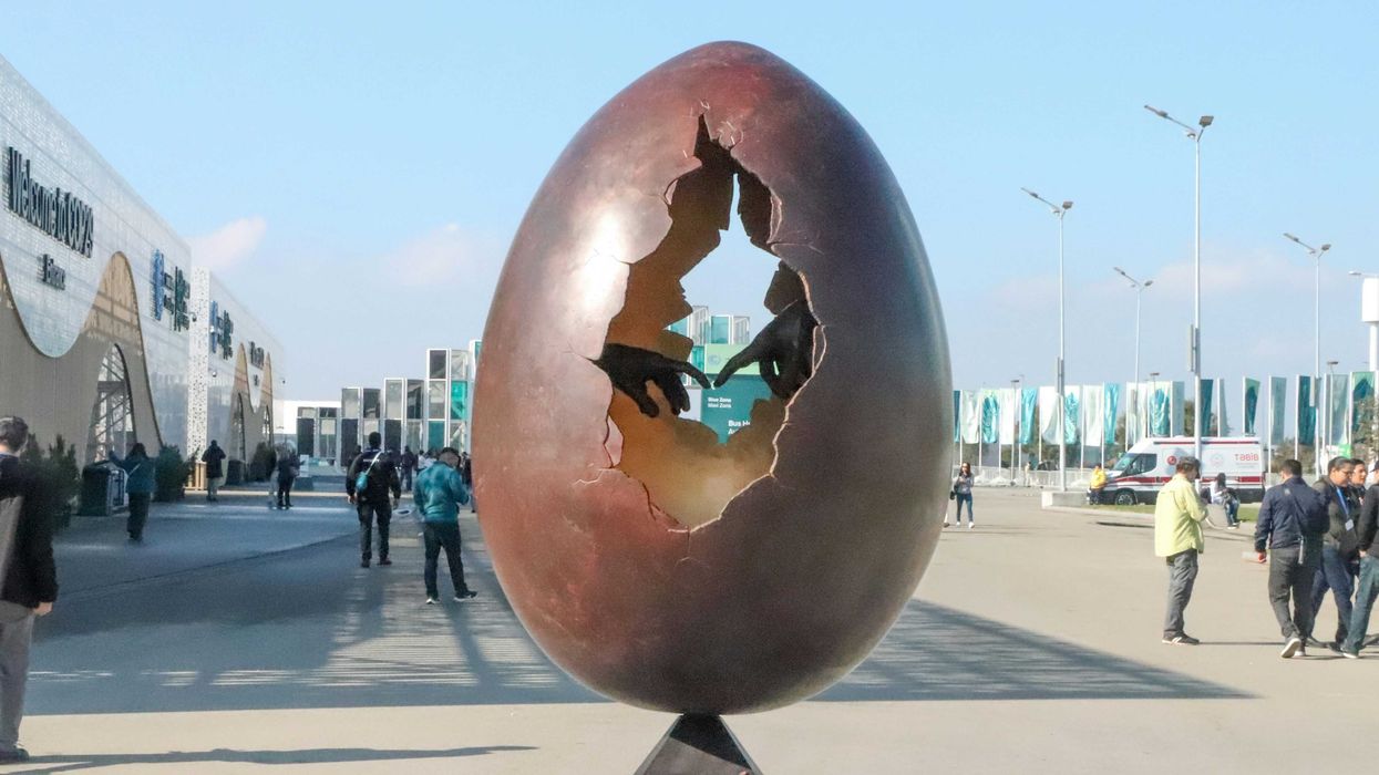 A large egg sculpture with a hole in the middle showing two hands with fingers touching, sitting on top of a metal pole.