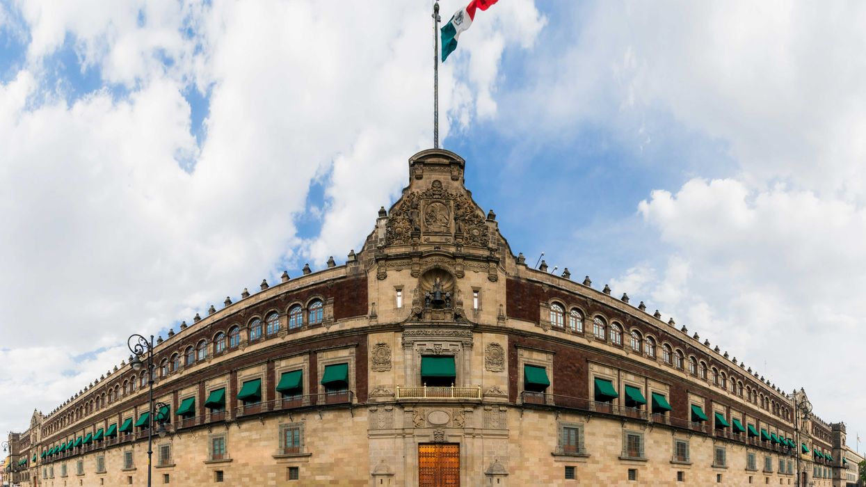 a large stone building with flags on top with Mexico's National Palace in the background.