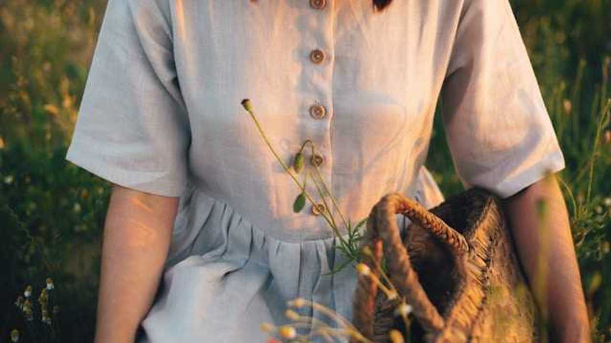 A linen dress on a woman in a field of wildflowers; she is holding a woven basket with plants inside
