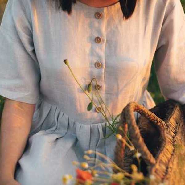A linen dress on a woman in a field of wildflowers; she is holding a woven basket with plants inside