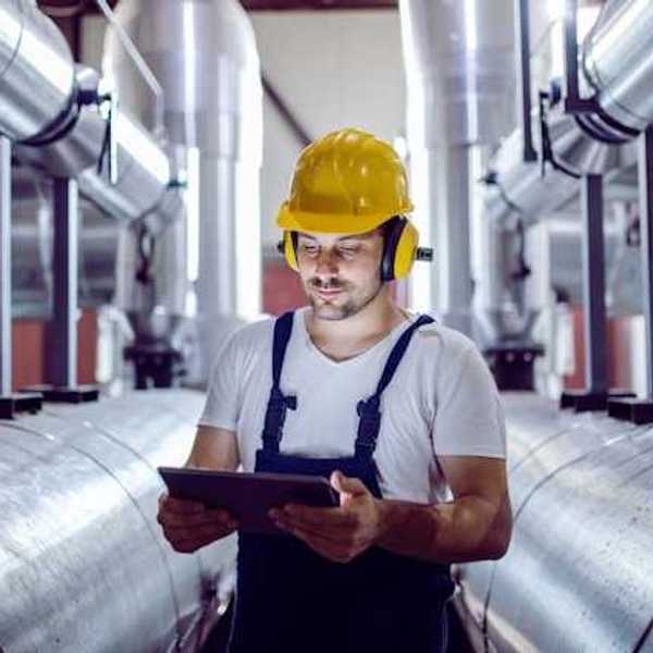A maintenance worker looking at a tablet while standing next to giant silver pipes