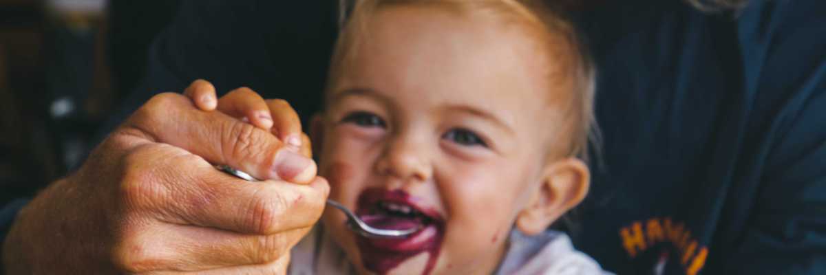 A man feeds a smiling toddler a purple gooey liquid from a spoon.