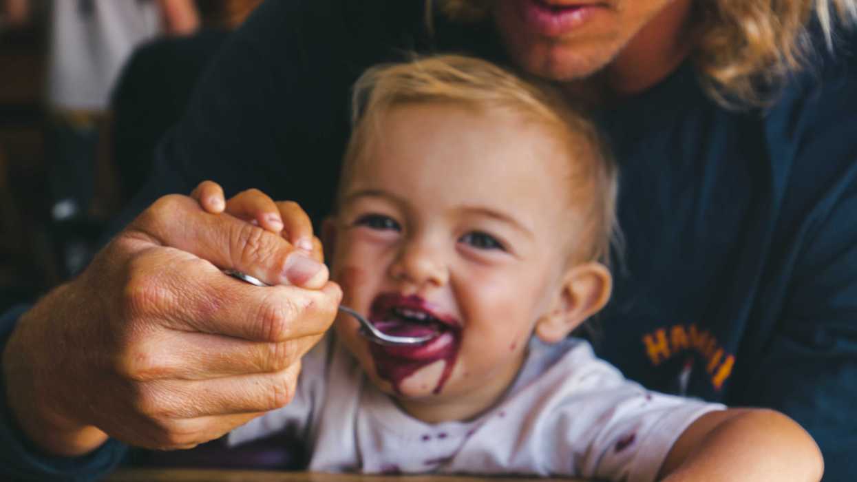 A man feeds a smiling toddler a purple gooey liquid from a spoon.