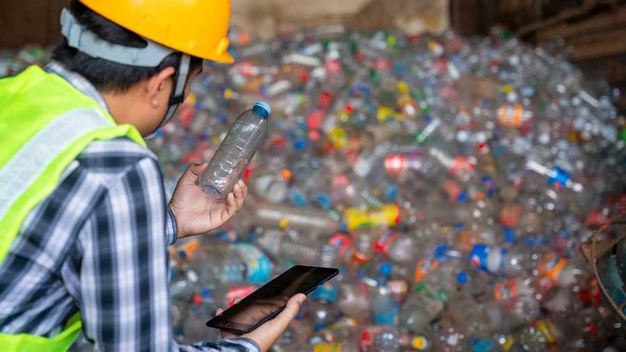 A man in a hard hat and neon vest, analyzing a plastic bottle in front of a large pile of them