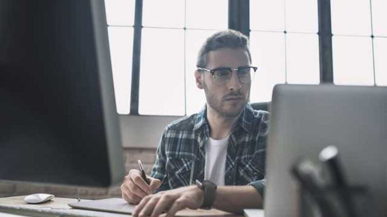 A man in a plaid shirt sitting at a desk in front of a laptop and monitor