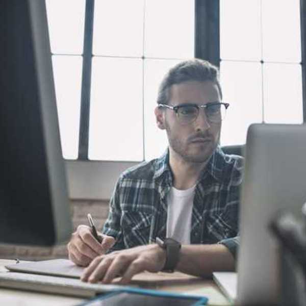 A man in a plaid shirt sitting at a desk in front of a laptop and monitor