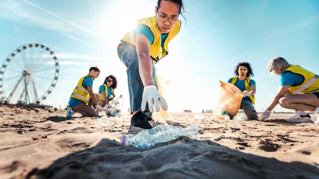 A man reaches for a discarded plastic bottle as 5 people wearing yellow vests pick up beach litter
