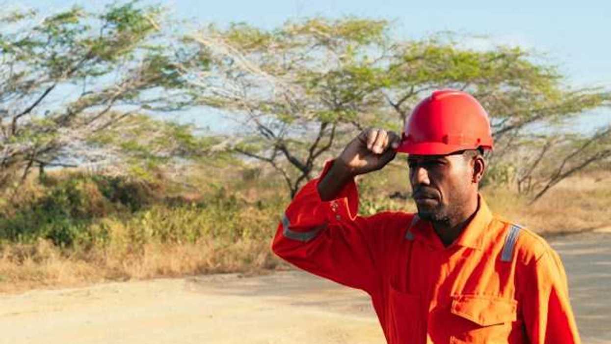A man wearing an orange work jumpsuit and hard hat walks down a dirt road