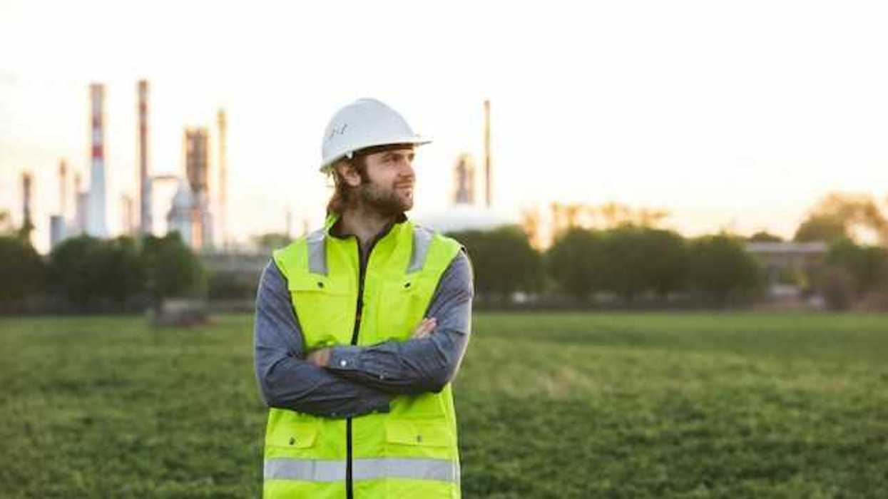 A man with a hard hat and neon vest on, posing in front of an oil refinery