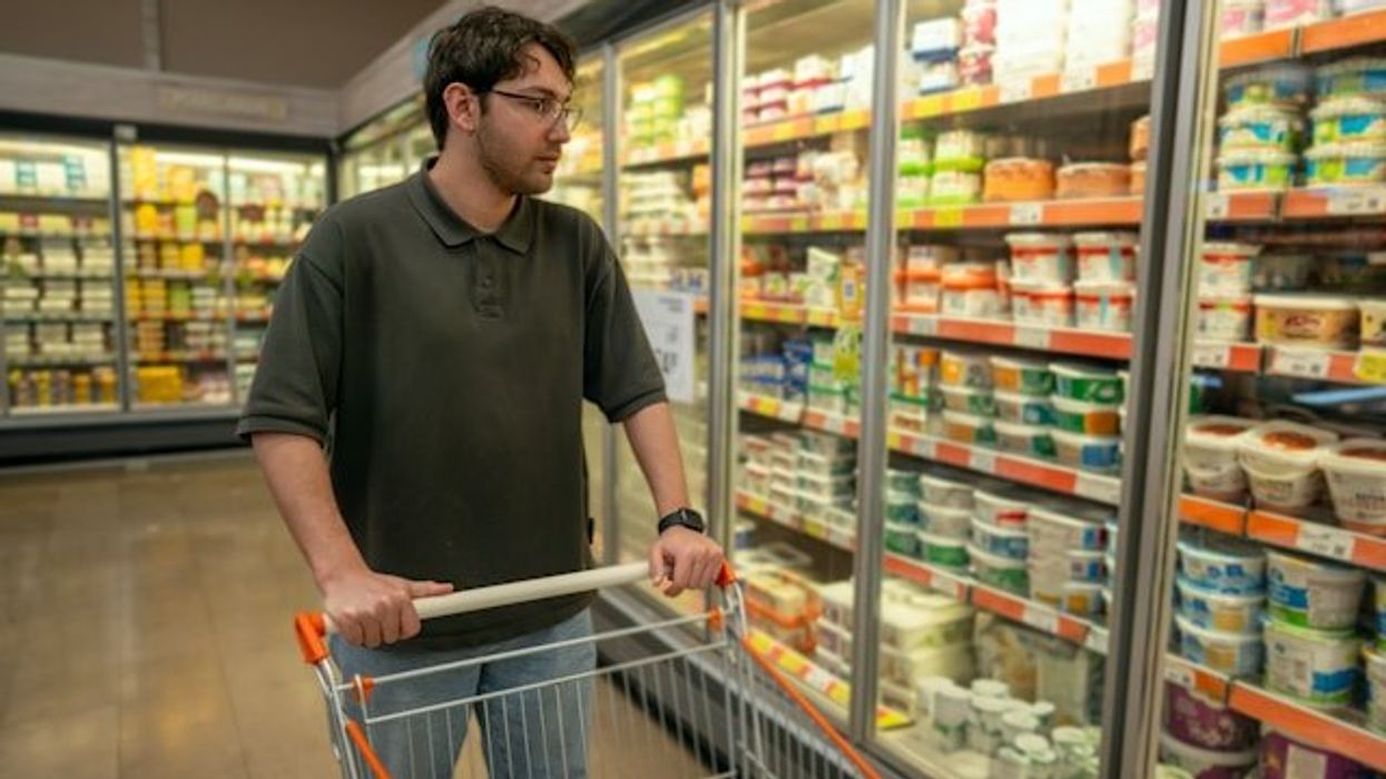 A man with a shopping cart looking at food in a grocery store refrigerator.