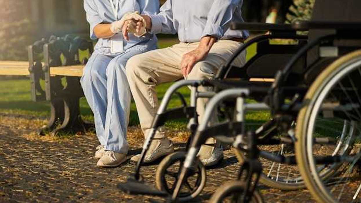 A nurse sits with a man on a bench with a wheelchair in the foreground