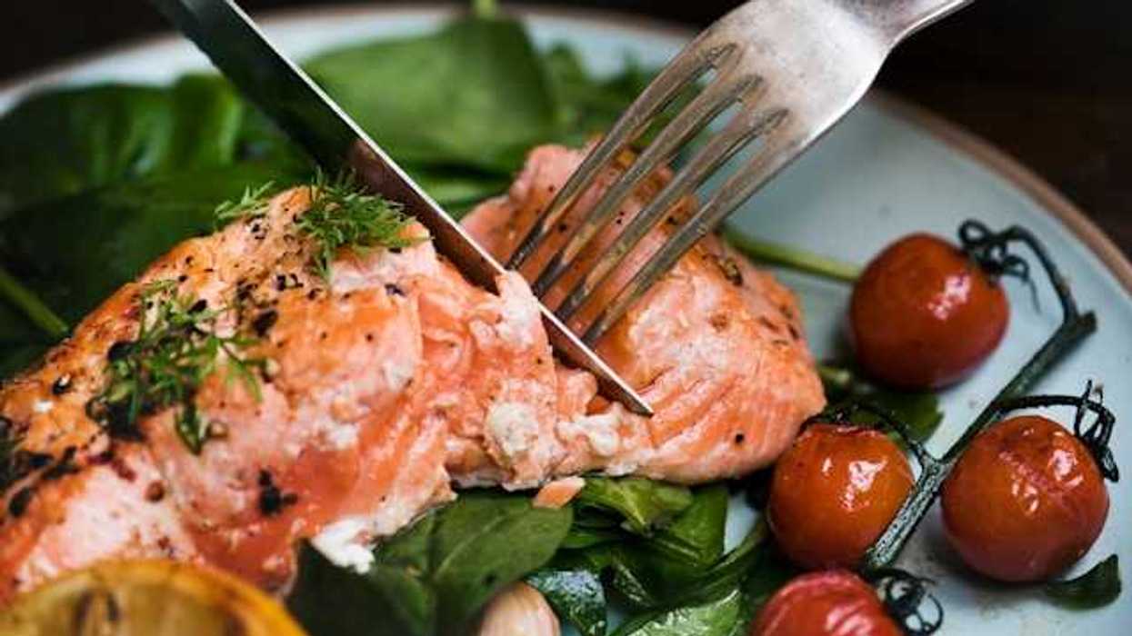A person cutting a salmon filet on a plate with spinach and cherry tomatoes