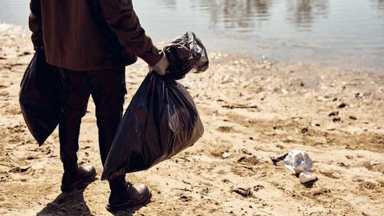 A person holding two plastic bags on a beach littered with trash