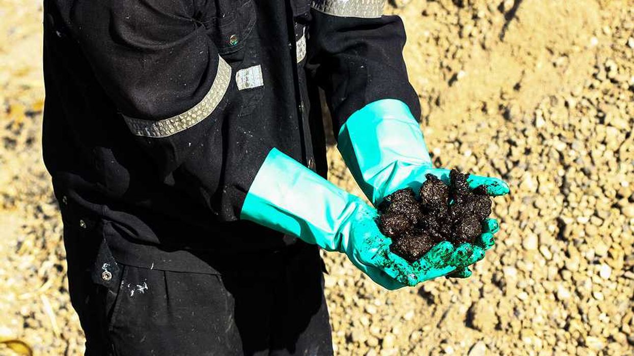A person in black protective clothing and rubber gloves holding black oily material